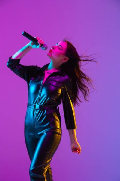 Caucasian female singer portrait isolated on purple studio background in neon light. Beautiful female model in black wear with microphone. Concept of human emotions, facial expression, ad, music, art.